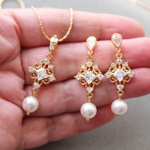 May include: Gold necklace and earrings set with a square-shaped pendant and matching earrings. The pendant and earrings feature a clear crystal stone surrounded by smaller crystals and a pearl drop.