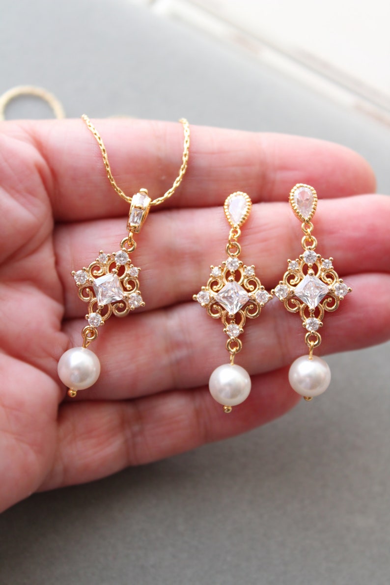 May include: Gold necklace and earrings set with a square-shaped pendant and matching earrings. The pendant and earrings feature a clear crystal stone surrounded by smaller crystals and a pearl drop.