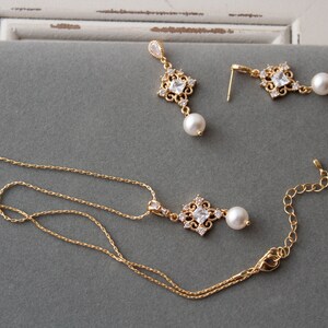 May include: A gold necklace with a pendant featuring a square-shaped crystal surrounded by smaller crystals and a pearl drop. Matching earrings with a similar design and pearl drops are also included.
