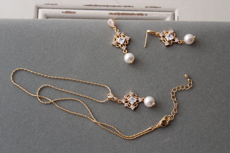 May include: A gold necklace with a pendant featuring a square-shaped crystal surrounded by smaller crystals and a pearl drop. Matching earrings with a similar design and pearl drops are also included.
