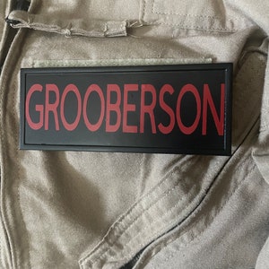 May include: A black rectangular patch with the name "GROOBERSON" in red lettering. The patch is attached to a tan jumpsuit, with a zipper and seams visible. The image suggests a costume or uniform.