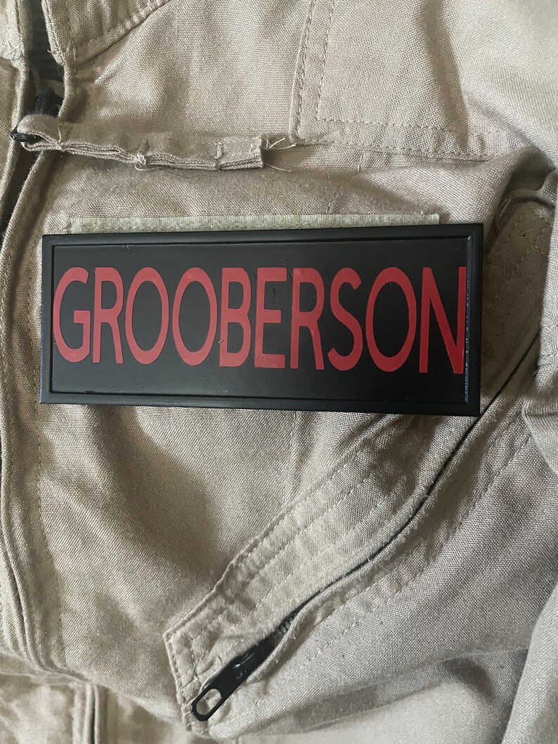 May include: A black rectangular patch with the name "GROOBERSON" in red lettering. The patch is attached to a tan jumpsuit, with a zipper and seams visible. The image suggests a costume or uniform.