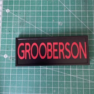 May include: A black name tag with the name "GROOBERSON" in red lettering. The tag is rectangular and set against a green grid background.