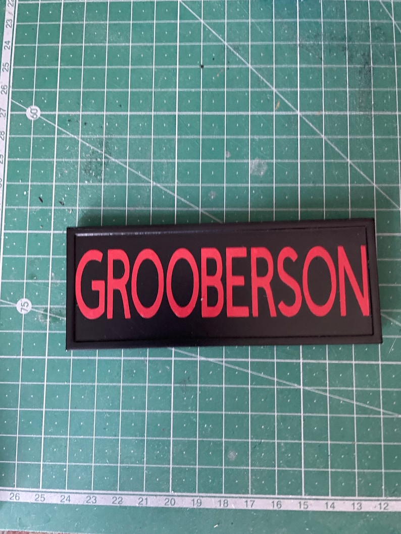 May include: A black name tag with the name "GROOBERSON" in red lettering. The tag is rectangular and set against a green grid background.
