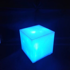 Op de afbeelding: Een blauwe kubusvormige lamp met een getextureerd oppervlak. De lamp is aan en straalt een helder blauw licht uit.