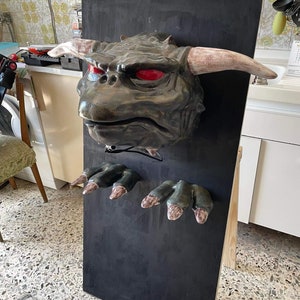 May include: A green and brown monster head with red eyes and horns mounted on a black board. The monster has two green hands with claws attached to the board.