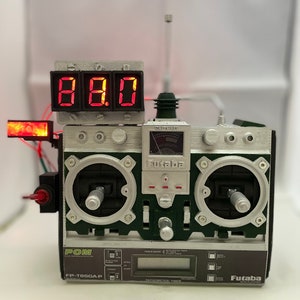 May include: A green and silver Futaba FP-T8SG-A-P radio control transmitter with a red digital display showing 8.8.0. The transmitter has two joysticks, a variety of buttons and knobs, and a small LCD screen.