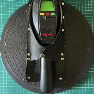 May include: A black leather holster with a black device with a green screen and red, yellow, and green lights. The holster is attached to a black circular base with a textured surface.