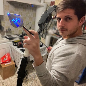 Puede incluir: Una persona sostiene un arco compuesto negro y gris con una luz azul en la flecha. La flecha tiene una punta blanca impresa en 3D con la palabra "PYM" en ella.