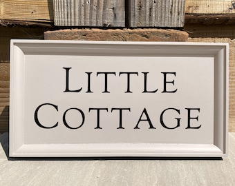 May include: A white wooden sign with a black frame and the text "LITTLE COTTAGE" in black lettering.