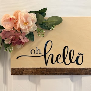 May include: Wooden sign with the words "oh, hello" in black script. The sign is decorated with faux flowers in shades of pink and peach, and green leaves. A dark brown wooden accent is at the bottom.