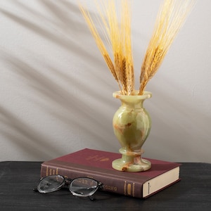 May include: A green onyx vase with a slender neck and a wide base, filled with dried wheat stalks. The vase is sitting on top of a burgundy book with the text "The Order of the Ring" visible.