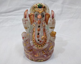 May include: A white crystal Ganesha statue with gold and red accents. The statue is sitting on a pink and gold base.