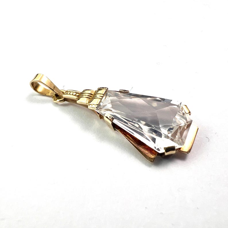 May include: A gold pendant with a large, clear, rectangular gemstone. The pendant has a detailed gold setting and a loop for a chain. The gemstone is faceted, catching the light and creating a sparkling effect.