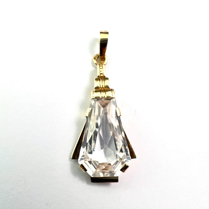 May include: A gold-colored pendant with a clear, faceted, teardrop-shaped crystal. The pendant has a rectangular top loop and geometric accents. The crystal is approximately 1.5 inches long.