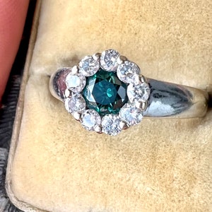 May include: A silver ring featuring a central teal gemstone surrounded by a halo of smaller, clear gemstones. The ring is set against a beige background, showcasing the intricate details of the jewelry.
