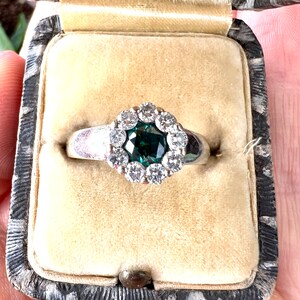 May include: A silver ring with a dark teal square-cut center stone, surrounded by a halo of small, round, clear stones. The ring is displayed in a vintage, velvet-lined jewelry box.