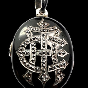 May include: Oval black pendant with a silver bail. The pendant features a silver monogram design encrusted with small, sparkling stones. The design is centered on the pendant and is surrounded by a black, glossy surface. The pendant is suspended from a silver bail.