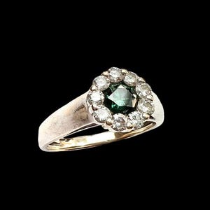 May include: A vintage ring with a central emerald-cut green gemstone, surrounded by a halo of smaller, clear gemstones. The ring has a silver band with a gold-colored interior, set against a black background.
