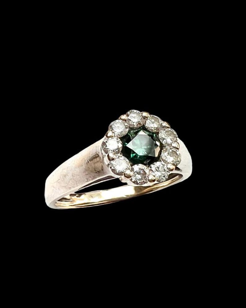 May include: A vintage ring with a central emerald-cut green gemstone, surrounded by a halo of smaller, clear gemstones. The ring has a silver band with a gold-colored interior, set against a black background.