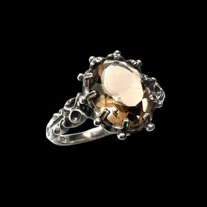 May include: A silver ring featuring a large, oval-cut, light brown gemstone set in a pronged mounting. The band and setting are crafted with intricate, raised metalwork details that extend from the base of the stone along the ring shank.
