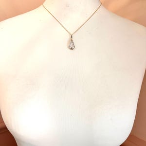 May include: A delicate gold necklace with a small, clear pendant. The pendant has a rectangular shape with a pointed bottom and a small, round accent. The necklace is displayed on a white mannequin.