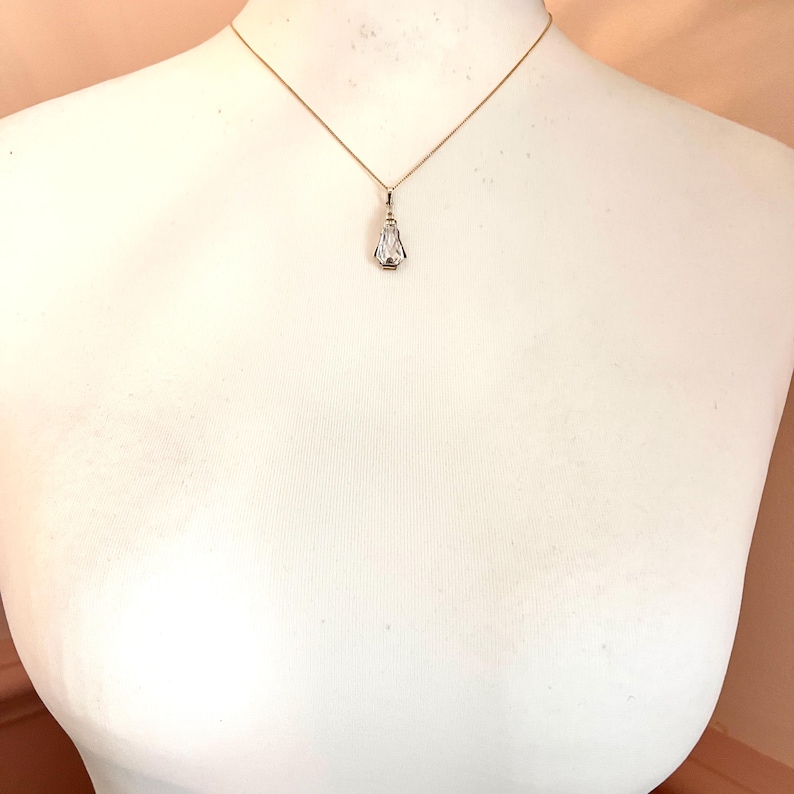 May include: A delicate gold necklace with a small, clear pendant. The pendant has a rectangular shape with a pointed bottom and a small, round accent. The necklace is displayed on a white mannequin.