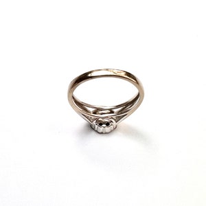 May include: A silver ring with a double band design. The ring features a small, round, possibly gemstone, center stone. The band has a split design near the center stone, creating an open, airy look. The ring is set against a white background.