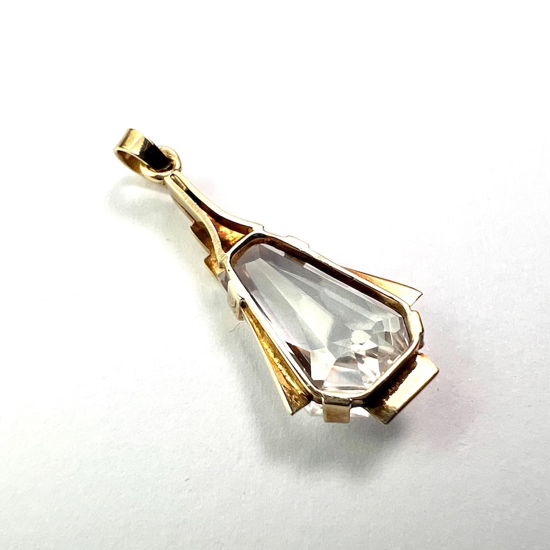 May include: A gold pendant with a clear, teardrop-shaped gemstone. The pendant has a rectangular bail and geometric gold accents around the stone. The gemstone is faceted, reflecting light.