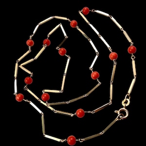 May include: A gold-toned necklace with alternating red beads and rectangular gold bars. The necklace is displayed against a black background, highlighting the contrast between the gold and red elements. The clasp is visible at the end of the necklace.