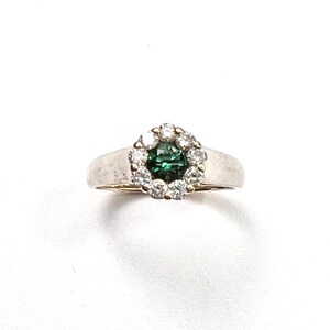 May include: A silver ring featuring a central teal gemstone surrounded by a halo of smaller, clear gemstones. The ring has a simple, polished band and a classic, elegant design, suitable for various occasions.