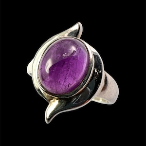 May include: A silver ring featuring a smooth, oval-shaped purple amethyst gemstone set in the centre. The metal band has a polished finish and curves around the stone with two pointed, wing-like extensions on the top and bottom sides.