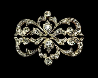 Könnte beinhalten: Aufwendige, vintage Brosche mit symmetrischem Design. Die Brosche hat eine silberfarbene Metalleinfassung, verziert mit zahlreichen klaren, funkelnden Edelsteinen. Das Design umfasst wirbelnde, floral inspirierte Elemente und einen zentralen, größeren Edelstein.
