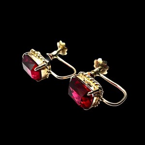 Może przedstawiać: Para zabytkowych kolczyków w kolorze złota z dużymi, fasetowanymi, prostokątnymi czerwonymi kamieniami szlachetnymi. Kolczyki mają zapięcie na śrubę i dekoracyjny element kwiatowy. Kamienie mają głęboki, bogaty czerwony kolor.
