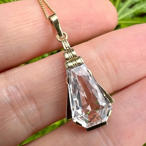 May include: A gold-toned pendant necklace featuring a large, clear, faceted rectangular gemstone. The pendant has a geometric design with a gold chain. The necklace is displayed against a blurred green background.
