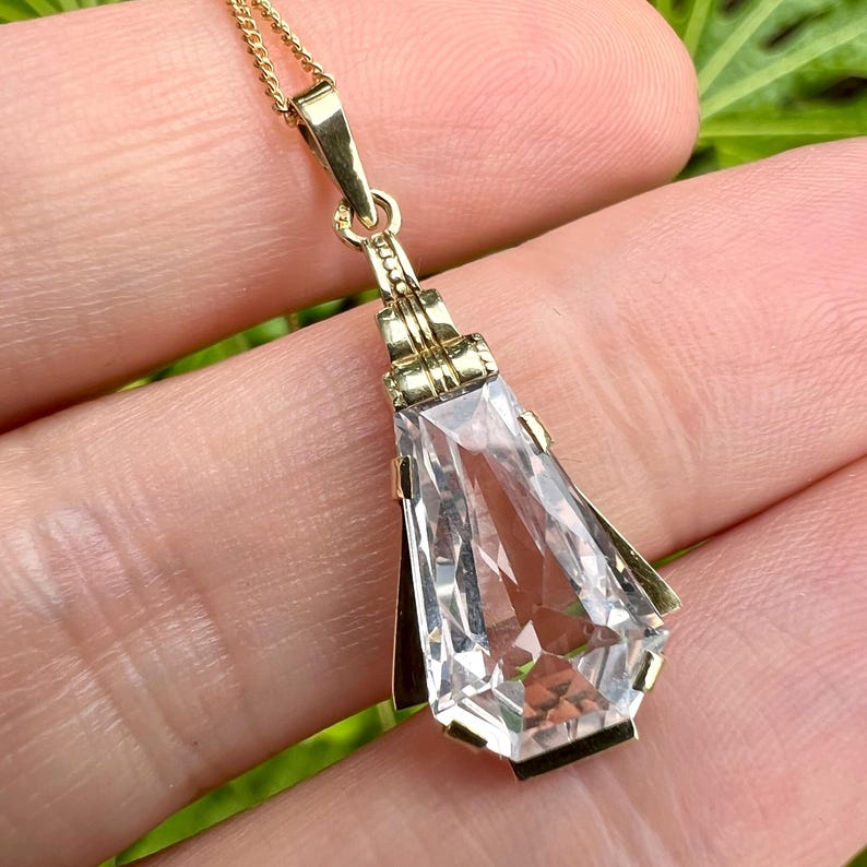 May include: A gold-toned pendant necklace featuring a large, clear, faceted rectangular gemstone. The pendant has a geometric design with a gold chain. The necklace is displayed against a blurred green background.