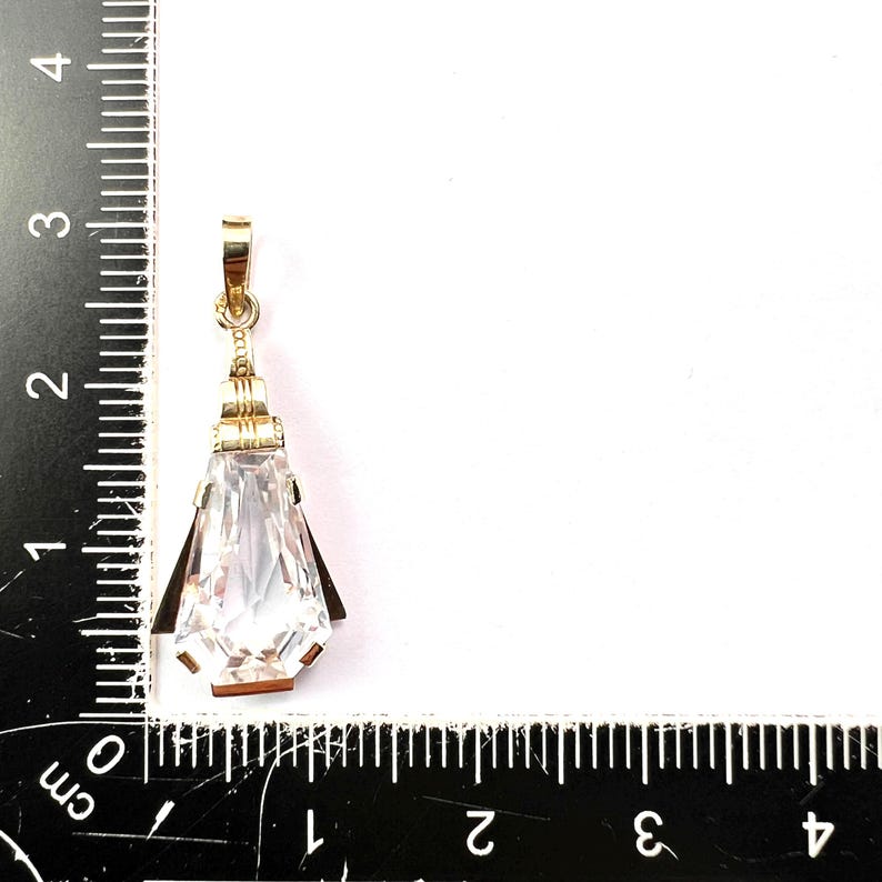 May include: A gold-colored pendant with a large, clear, faceted crystal. The pendant is teardrop-shaped, with a rectangular top and a pointed bottom. The crystal is approximately 1.5 inches long. The pendant is on a white background.
