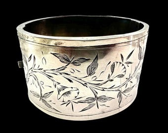 May include: A silver-toned, cylindrical bracelet with intricate floral engravings. The design features detailed leaves and flowers, creating a delicate pattern around the band. The bracelet has a clasp closure and a polished finish.