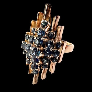 Könnte beinhalten: Ein goldfarbener Ring mit einer Ansammlung dunkelblauer Edelsteine. Der Ring hat ein abstraktes Design mit vertikalen und abgewinkelten Goldstäben, die die Edelsteine umgeben, wodurch ein einzigartiges, auffälliges Schmuckstück entsteht.