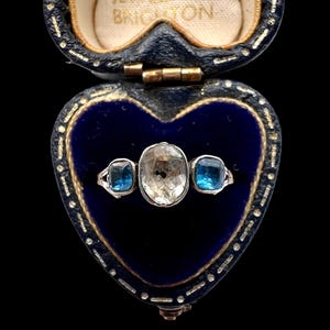 May include: An antique silver ring with an oval-cut, light-colored center stone and two square, blue side stones. The ring is displayed in a heart-shaped, blue velvet jewelry box with gold trim. The box is open, revealing the ring.