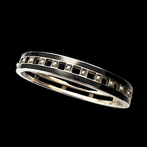 Pode incluir: Uma pulseira de bracelete de prata com uma faixa de esmalte preto. A pulseira apresenta uma série de pequenos recortes quadrados preenchidos com pequenas pérolas brancas redondas. A pulseira está em um fundo preto.