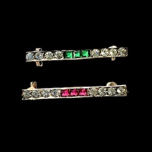 May include: Two bar brooches featuring a metallic frame set with clear round rhinestones. The top brooch includes three square-cut green stones in the centre, while the bottom brooch features three square-cut red stones in the centre.