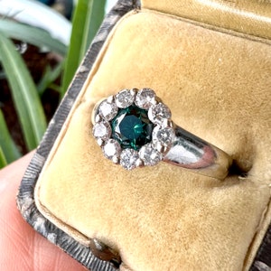 May include: A silver ring featuring a dark teal gemstone surrounded by a halo of small, clear gemstones. The ring is set in a vintage-style setting and is displayed in a velvet-lined jewelry box.