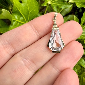 May include: A gold pendant with a clear, faceted crystal. The pendant has a rectangular shape with a pointed bottom and is held by a gold bail. The crystal is clear and reflects light. The pendant is held in a hand against a background of green leaves.