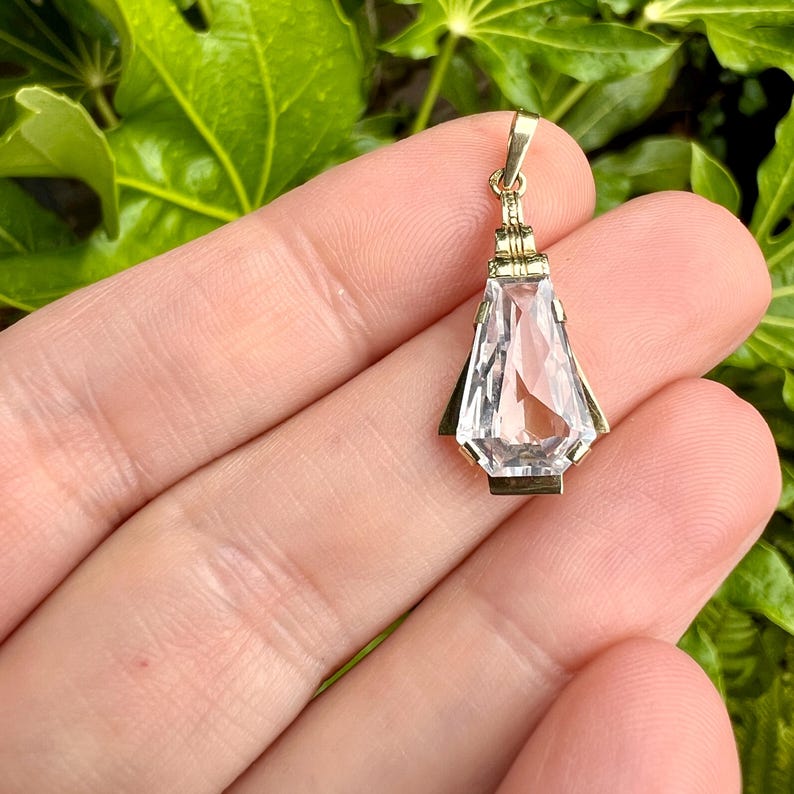 May include: A gold pendant with a clear, faceted crystal. The pendant has a rectangular shape with a pointed bottom and is held by a gold bail. The crystal is clear and reflects light. The pendant is held in a hand against a background of green leaves.