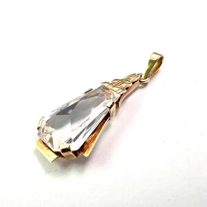 May include: A gold pendant featuring a large, clear, rectangular gemstone. The gemstone is faceted, catching the light. The pendant has a decorative gold setting and a loop for a chain. The design is elegant and classic.