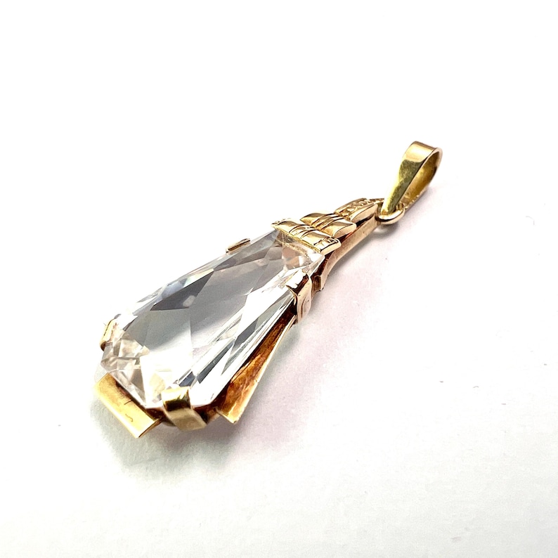 May include: A gold pendant featuring a large, clear, rectangular gemstone. The gemstone is faceted, catching the light. The pendant has a decorative gold setting and a loop for a chain. The design is elegant and classic.
