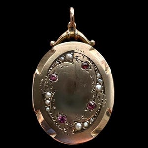 May include: An oval gold locket pendant featuring an engraved floral wreath motif. The design is accented with small white pearls and deep red gemstones arranged in a circular pattern. The locket includes a decorative gold bail at the top.