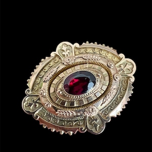 Op de afbeelding: Een ovale gouden broche met een centrale dieprode gefacetteerde edelsteen. Het metalen frame is versierd met ingewikkelde gegraveerde patronen, waaronder korenaren en bloemmotieven, die de centrale steen omringen met een getextureerde rand.