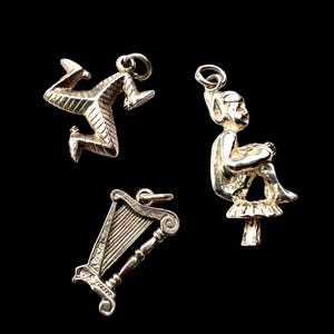May include: Three silver charms against a black backdrop. One depicts a three-legged symbol, another an elf seated on a mushroom, and the third a harp. These charms are likely for a necklace or bracelet.